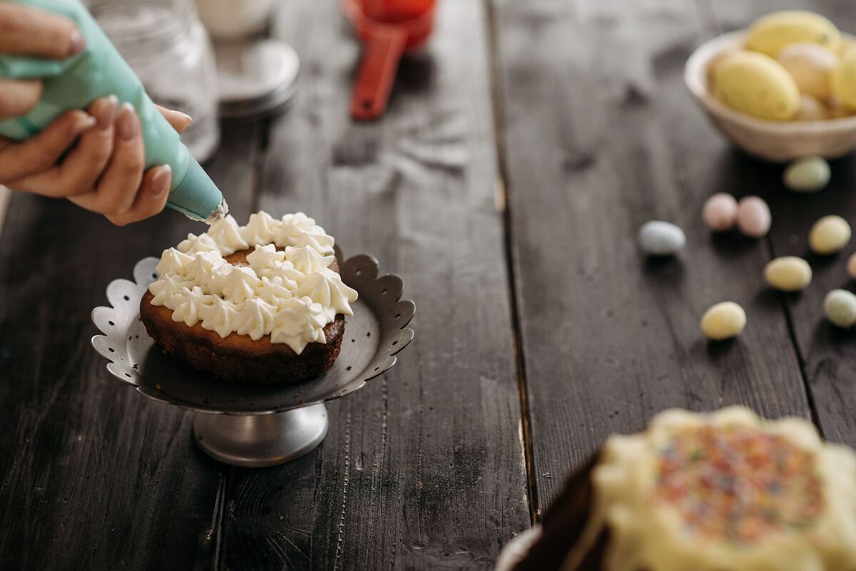 A piping bag laying swirls of cream onto a small cake on a dark wooden table