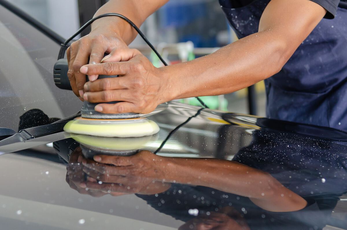 A detailer's hands guiding a dual-action polisher across the bonnet of a dark car.