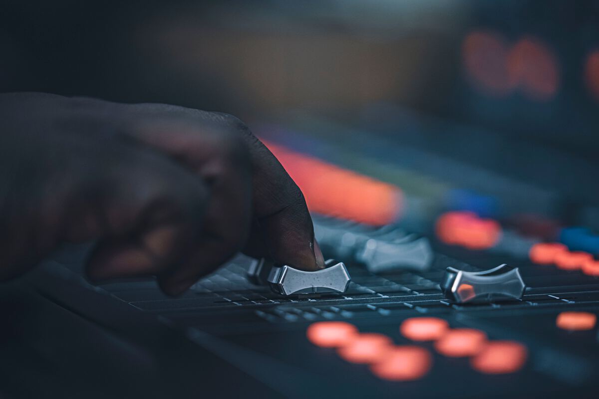 A hand adjusting the faders on an audio mixing console in a dark studio