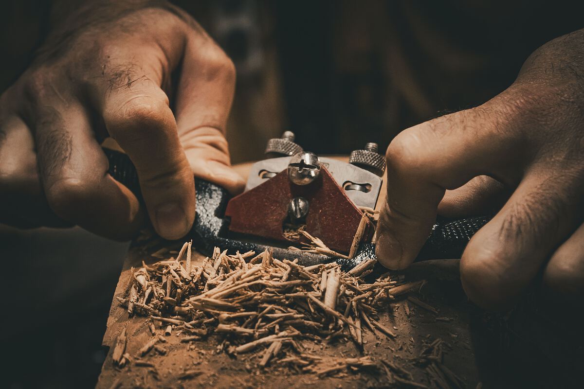 A woodworker's hands pushing a hand plane along a board, curls of shaving falling away.